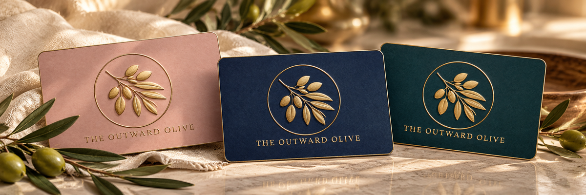The Outward Olive gift cards in pink, navy and teal with the gold olive branch logo, on a marble tabletop with olive branches